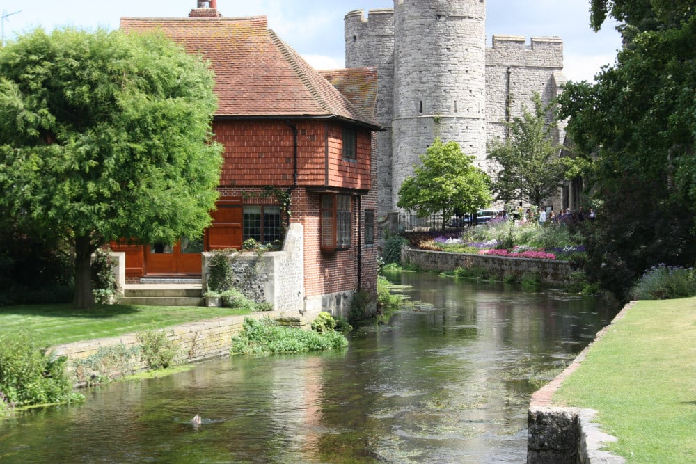Picture of a river with a building on the side of the water and another older tower like building further. A photograph of Westgate Gardens in Canterbury by See Kent
