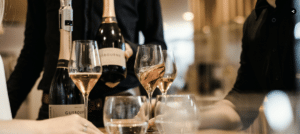 Wine glasses on a table with hands on the table and a person holding a bottle of wine to show them.