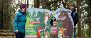 A board of Gruffalo and other images for the party trail and children in a hole in the board and a man and a woman in jackets and hats standing each side of the board with trees behind. For for Christmas in Kent ideas for Tunbridge Wells round-up on the website seekent.co.uk and the Gruffalo Party Trail at Bedgebury Pinetum