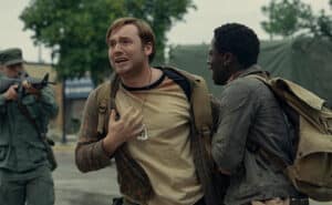 Two men with a man in military outfit aiming gun at them for New film releases for cinemas in Kent and The Long Walk from Lionsgate