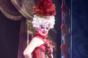 Person with huge white wig with a red hat on top with pantomime like make up for Graphic of a white and black cow for the Pantomime in Kent guide for 2025-6