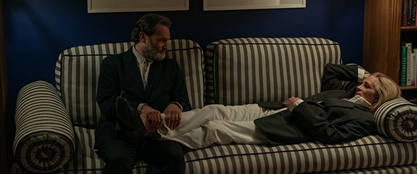 Woman lying on a striped sofa with her feet on a man's lap sitting at the end. For new film After The Hunt from Sony Pictures