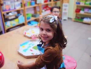 Young girl smiling at a table with crafts and a child in the background for Adult and two children in woods by Siim Lukka at -unsplash for 30 Things To Do in Kent for October Half Term guide